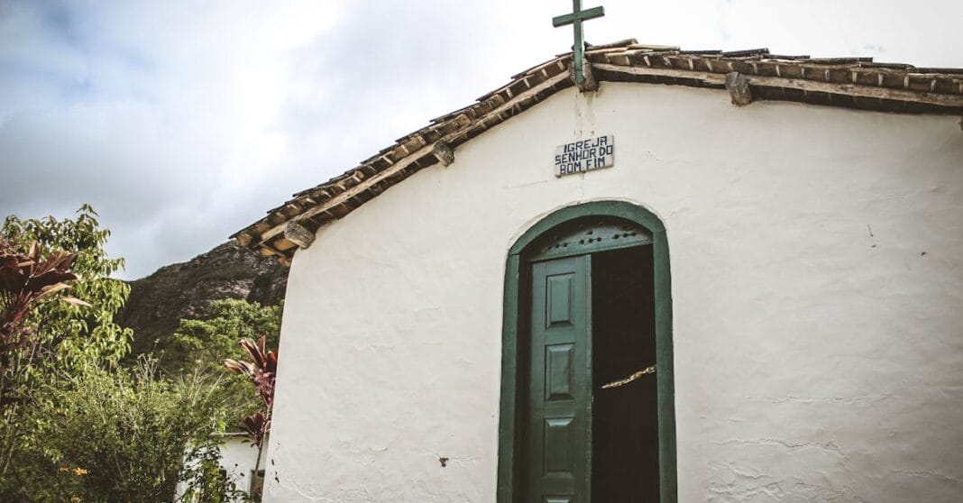 Capela histórica em Mocugê, Brasil, com porta verde e cruz de madeira, rodeada de vegetação nativa.
