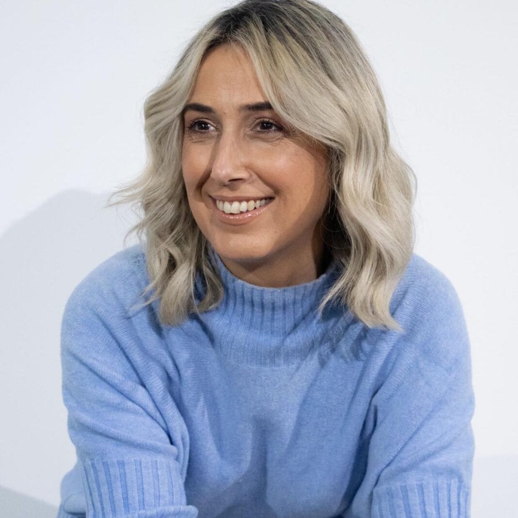 Retrato de uma mulher adulta, de cabelo loiro ondulado, vestida com uma camisola azul-clara, sorridente, fotografada em ambiente interior.