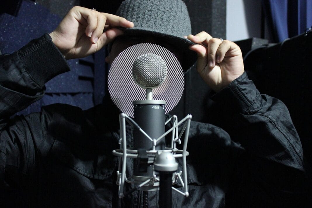 Homem num estúdio de gravação a ajustar o boné enquanto fala num microfone profissional, sugerindo o acto de captar áudio ou uma gravação confidencial.