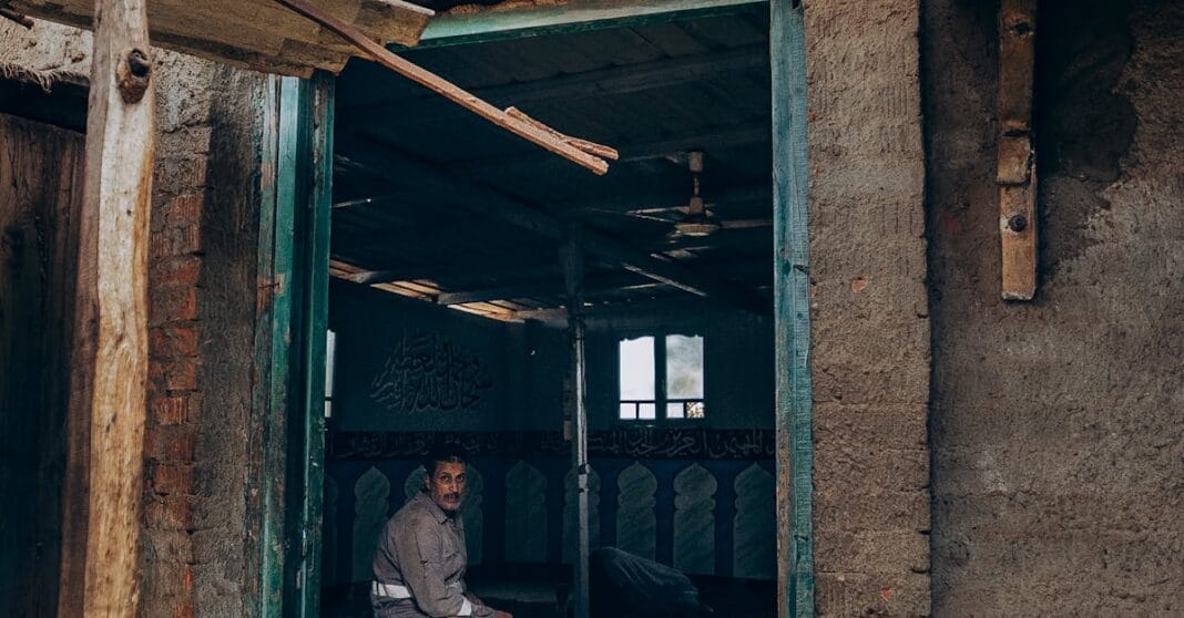 Interior de uma mesquita simples com um homem sentado junto à entrada, em ambiente desgastado.