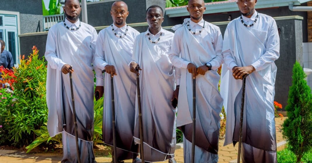 Homens vestidos com trajes cerimoniais tradicionais posam alinhados num pátio exterior, segurando cajados e usando colares rituais.