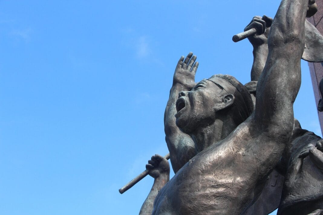 Escultura de figuras humanas em postura de resistência, braços erguidos sob um céu azul, simbolizando força e sobrevivência.