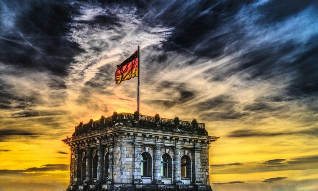 Fachada superior do Reichstag em Berlim com a bandeira da Alemanha ao vento, iluminada por um pôr do sol dramático com nuvens densas no céu.
