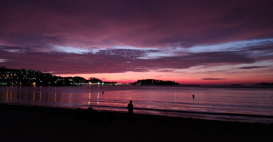 Pôr do sol sobre a baía de Baiona, com silhuetas na praia e luzes da cidade refletidas no mar.