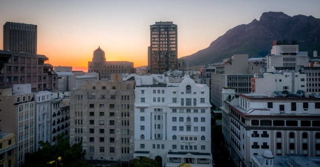 Vista urbana de Cidade do Cabo ao amanhecer, com edifícios históricos no primeiro plano e a Table Mountain ao fundo iluminada por luz suave.