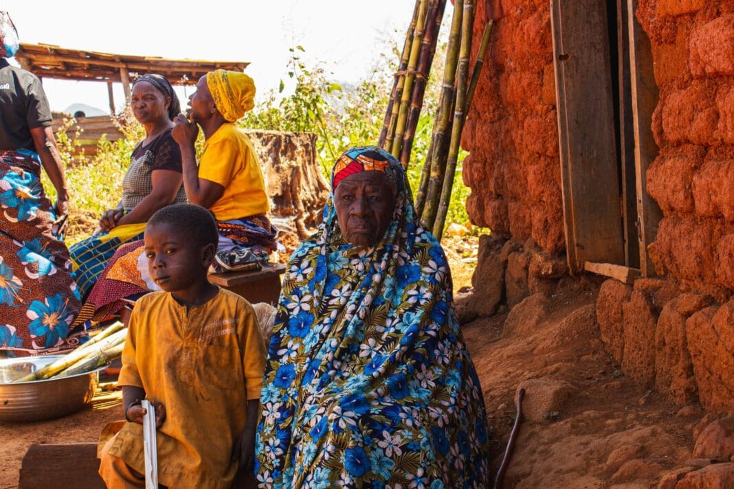 Mulher idosa e crianças sentadas à porta de uma casa de tijolo, numa aldeia da Nigéria