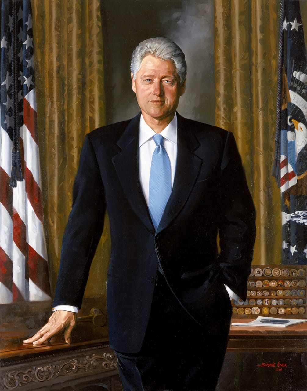 Retrato oficial de Bill Clinton de pé no Gabinete Oval, com fato escuro e gravata azul, diante da secretária presidencial e das bandeiras dos Estados Unidos.