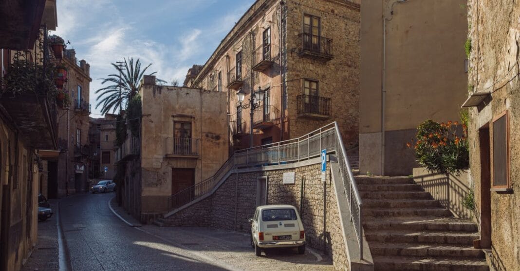 Rua ensolarada em Caccamo, Sicília, com edifícios históricos e carro estacionado.