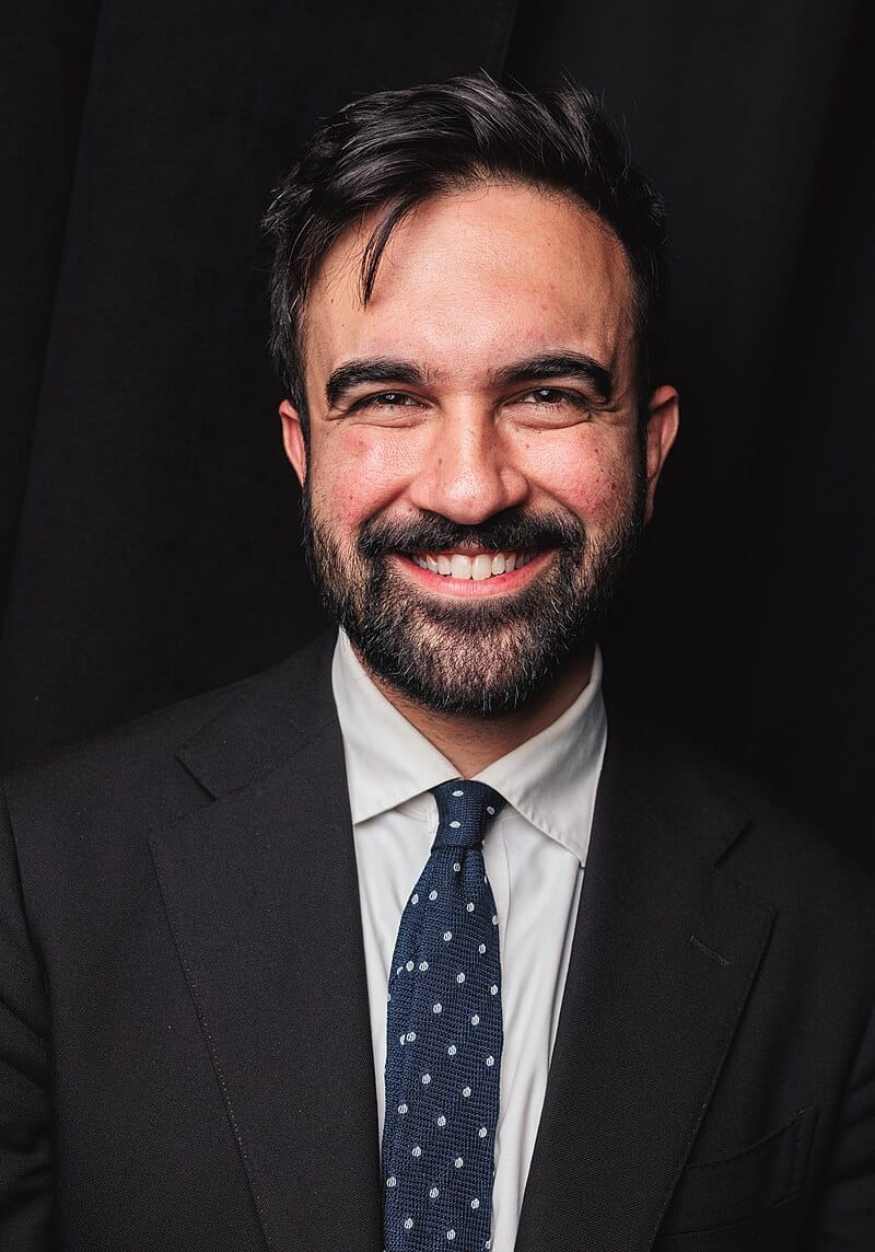 Zohran Mamdani sorridente, com fato escuro e gravata azul com pontos brancos.