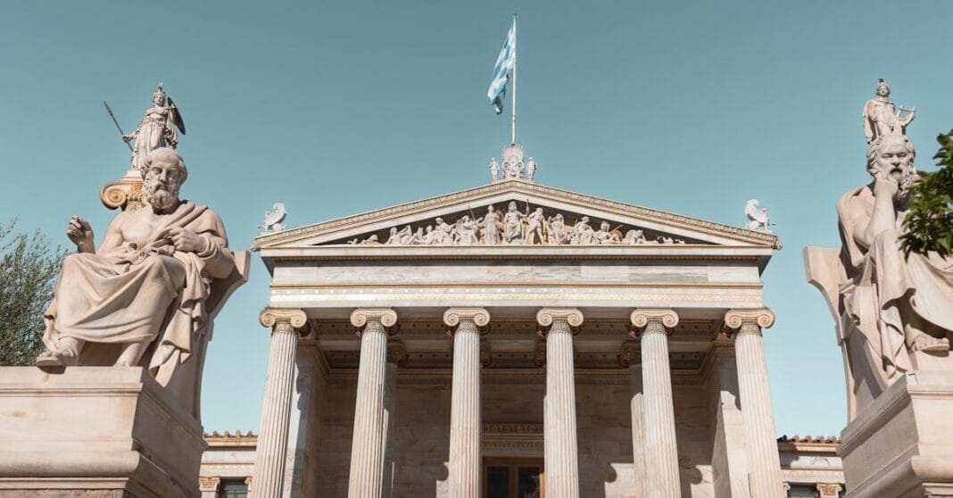 Fachada da Academia de Atenas com colunas clássicas, estátuas de filósofos sentados e a bandeira grega ao centro, sob um céu limpo.