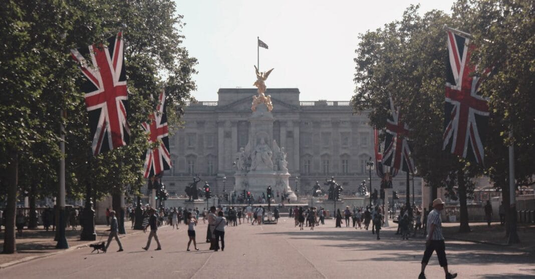 Vista de Buckingham Palace, em Londres, com bandeiras do Reino Unido e transeuntes num dia comum.