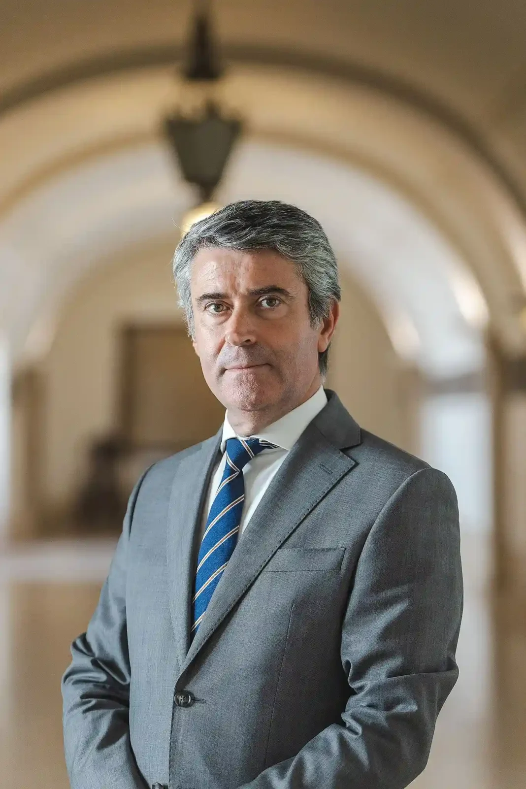 Retrato de José Luís Carneiro em pé, de fato cinzento e gravata às riscas, num corredor com arcos desfocados ao fundo.