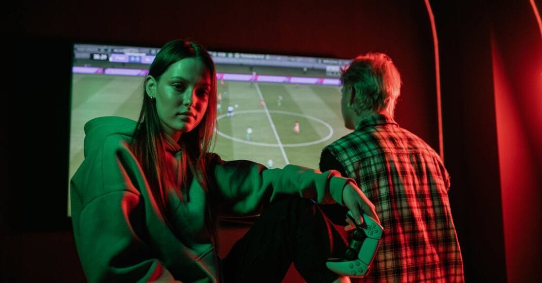 Dois jovens a jogar videojogo de futebol com comandos, iluminação vermelha e verde, ecrã com campo de futebol ao fundo