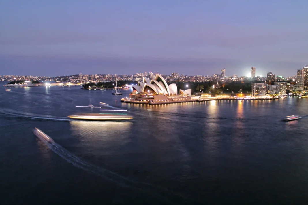 Vista nocturna do porto de Sydney, com embarcações e edifícios iluminados na margem.
