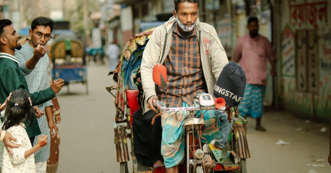 Homem empurra um carrinho carregado numa rua movimentada, com transeuntes ao fundo.