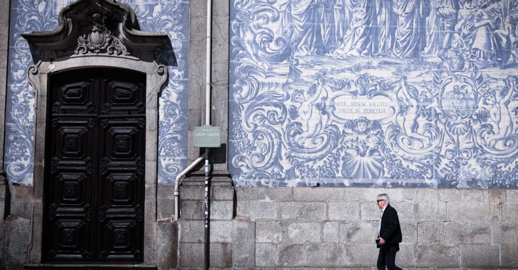 Homem caminha junto a uma parede de azulejos azuis tradicionais, numa rua urbana, com uma porta antiga ao fundo.