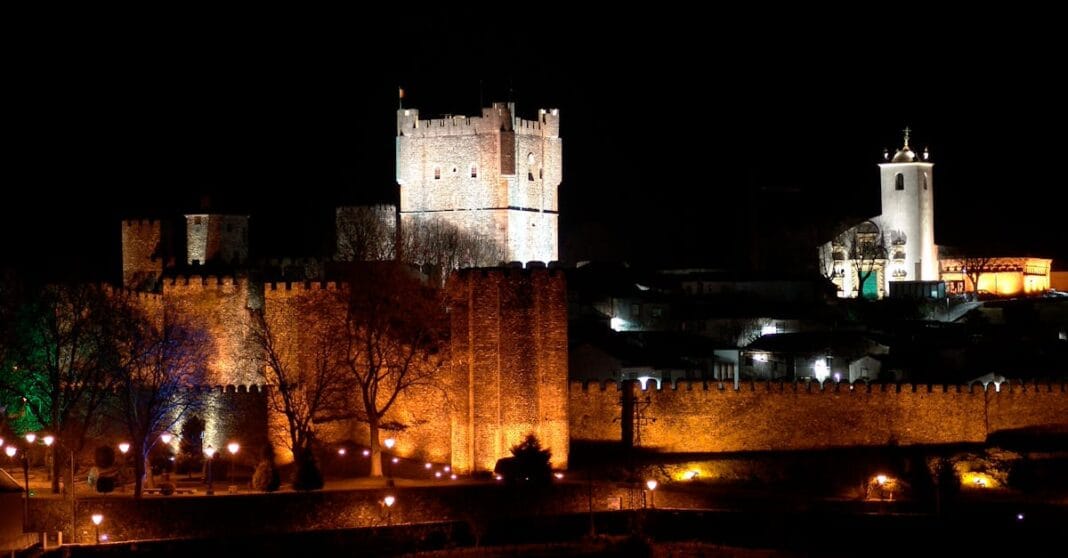 Castelo e muralhas iluminados à noite em Bragança, com torre principal ao centro e edifícios ao fundo.