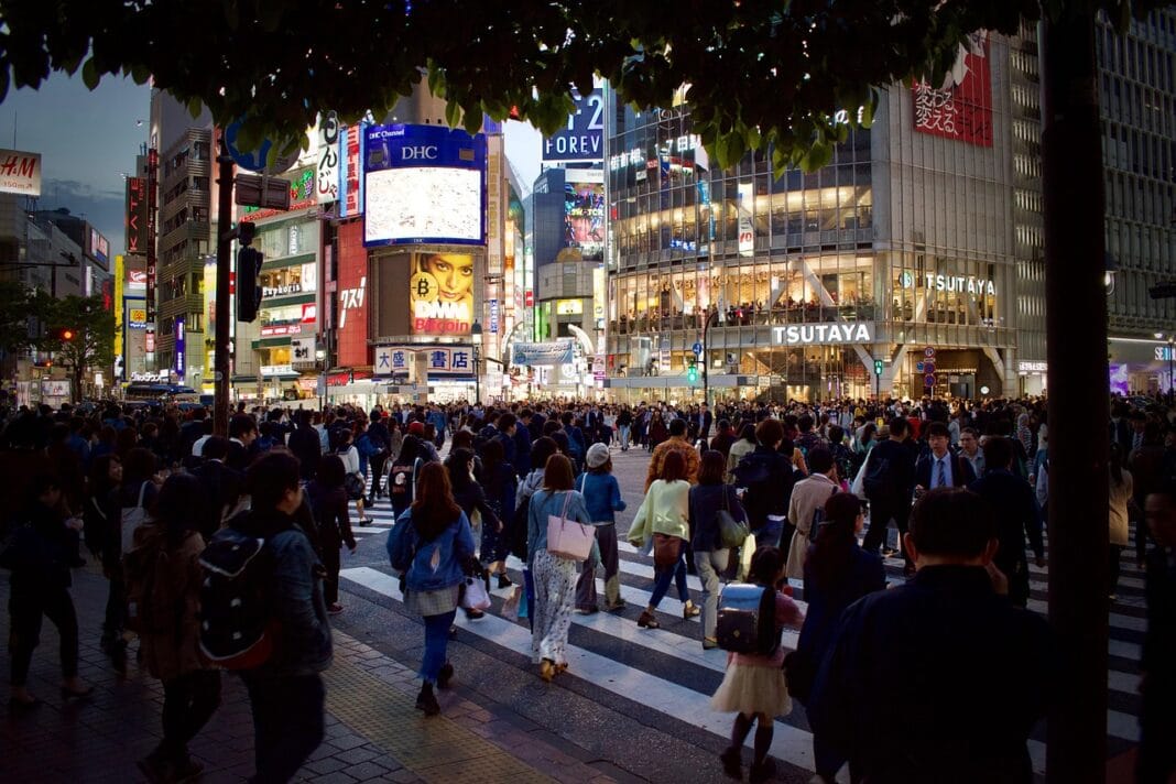 Multidão atravessa um cruzamento iluminado no centro de Tóquio ao início da noite, símbolo de uma cidade em movimento constante.