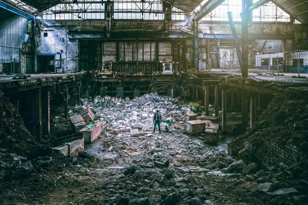 Interior de uma fábrica industrial abandonada, com maquinaria destruída e entulho acumulado no chão.