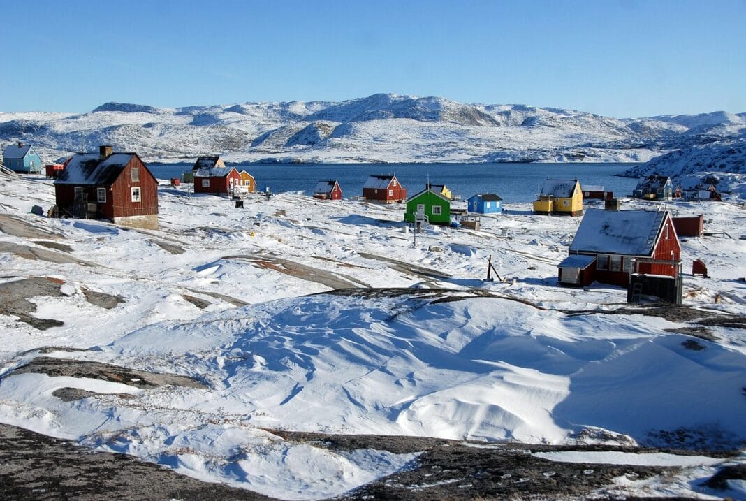 Casas coloridas numa aldeia costeira da Gronelândia, com neve, mar e montanhas ao fundo.