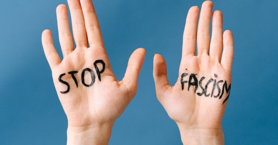Duas mãos erguidas com as palavras “STOP” e “FASCISM” escritas nas palmas, sobre fundo azul.