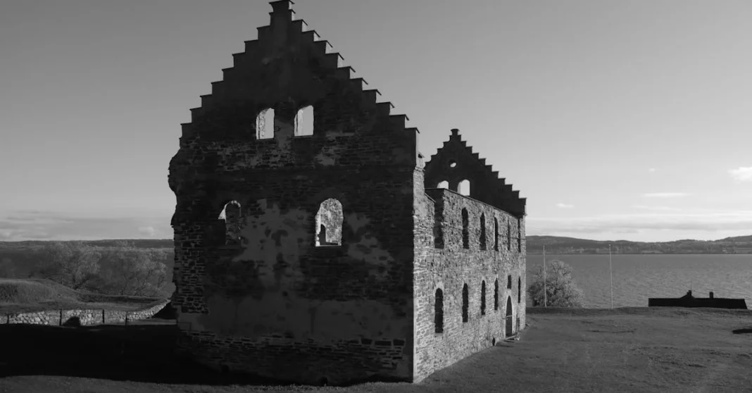 Ruínas de um edifício gótico em pedra, em fotografia a preto e branco, num campo junto a um lago.