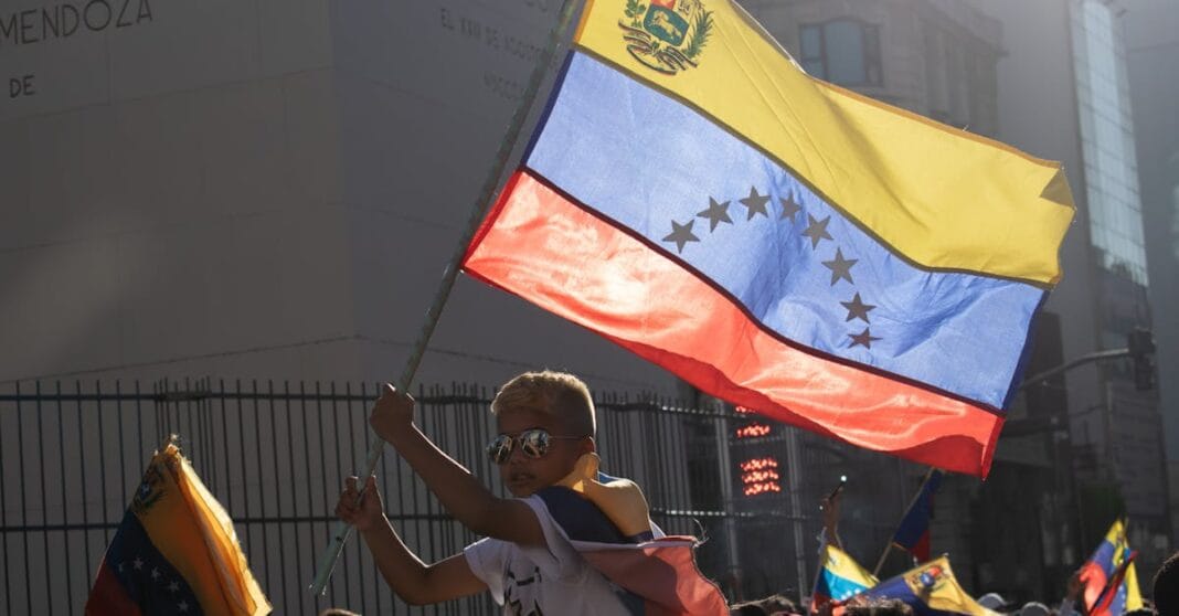 Pessoa com óculos de sol segura uma grande bandeira nacional, com faixas amarela, azul e vermelha e um arco de estrelas, num cenário urbano.