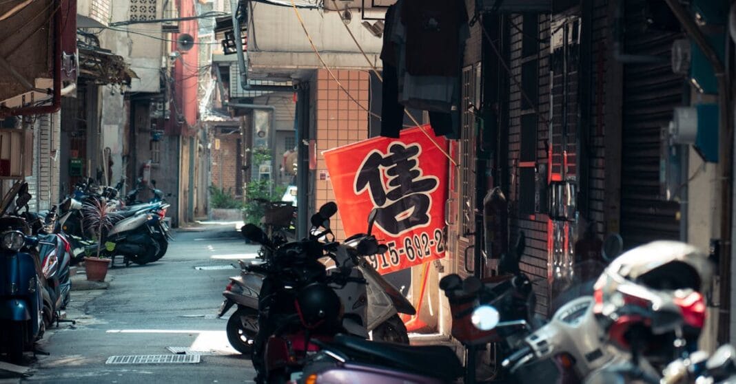 Rua estreita com várias motas estacionadas e uma pessoa em scooter; ao centro, uma bandeira/vermelha com um grande carácter chinês pendurada num suporte.