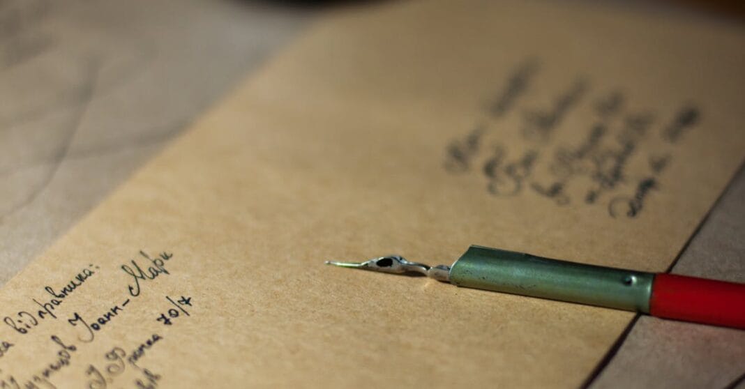 Caneta de aparo pousada sobre papel com escrita manuscrita, em tons quentes.