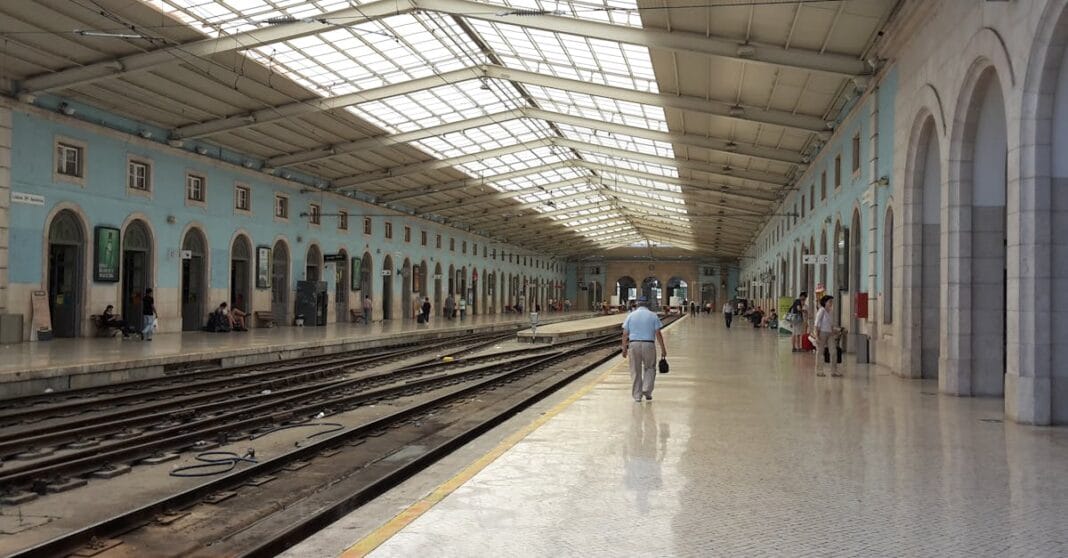 Interior de uma estação de caminho de ferro pintada de azul celeste, com passageiros a circular junto às linhas de comboio.