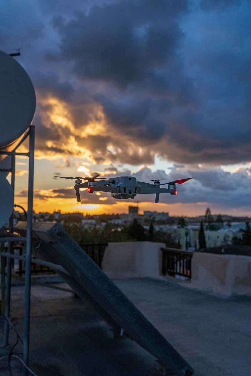Drone quadcopter em voo ao pôr-do-sol, com luzes vermelhas, sobre uma cidade desfocada.
