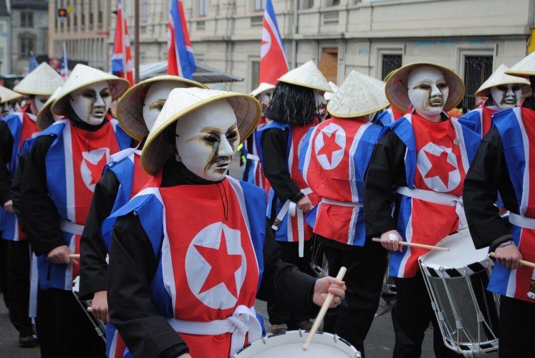 Grupo de pessoas com máscaras brancas e chapéus cónicos, em marcha na rua, com coletes vermelhos com um círculo branco e estrela, e um tambor ao lado.