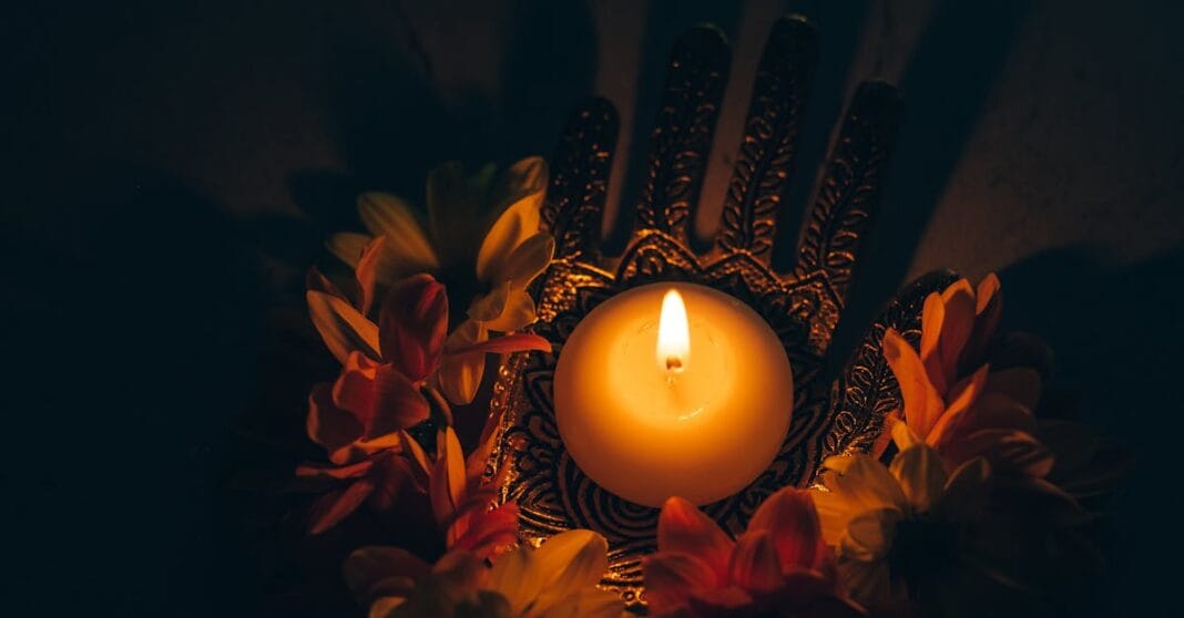 Vela acesa ao centro, em fundo escuro, rodeada por flores alaranjadas e elemento decorativo.