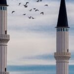 Two tall minarets stand elegantly against a blue sky with birds flying around.