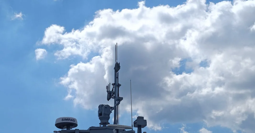 Navy ship mast with radar equipment beneath a clear blue sky with clouds.
