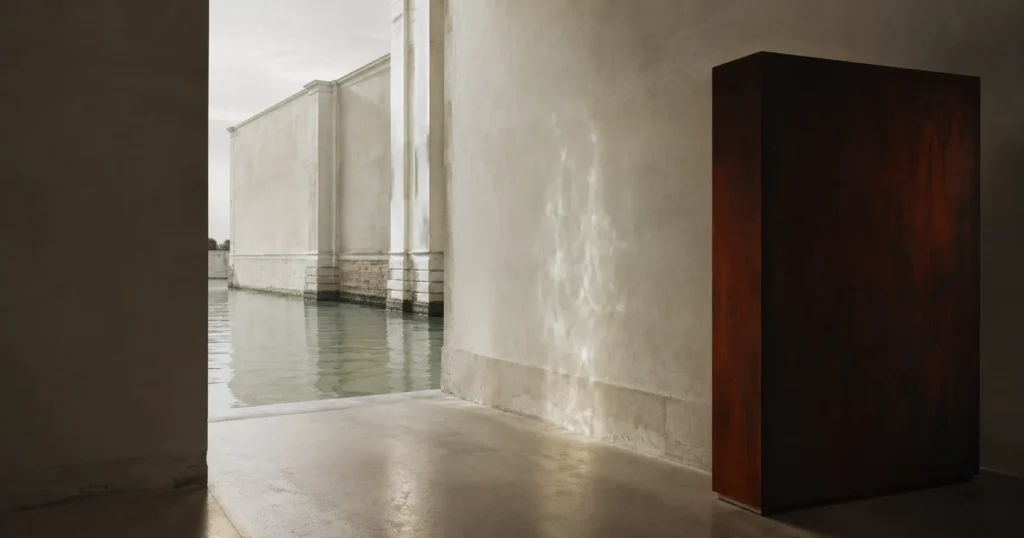 Imagem horizontal de uma passagem arquitetónica junto à água, com luz refletida nas paredes e um bloco escuro, associada à Bienal de Veneza 2026.