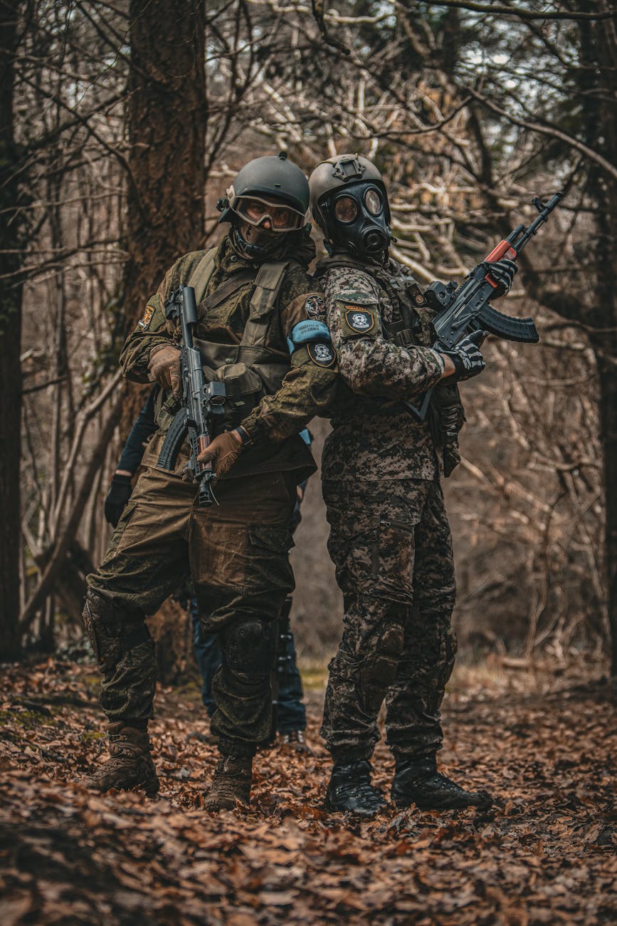 Dois combatentes em uniforme camuflado, armados e com capacetes, um deles usando máscara de gás, posicionados numa zona florestal densa