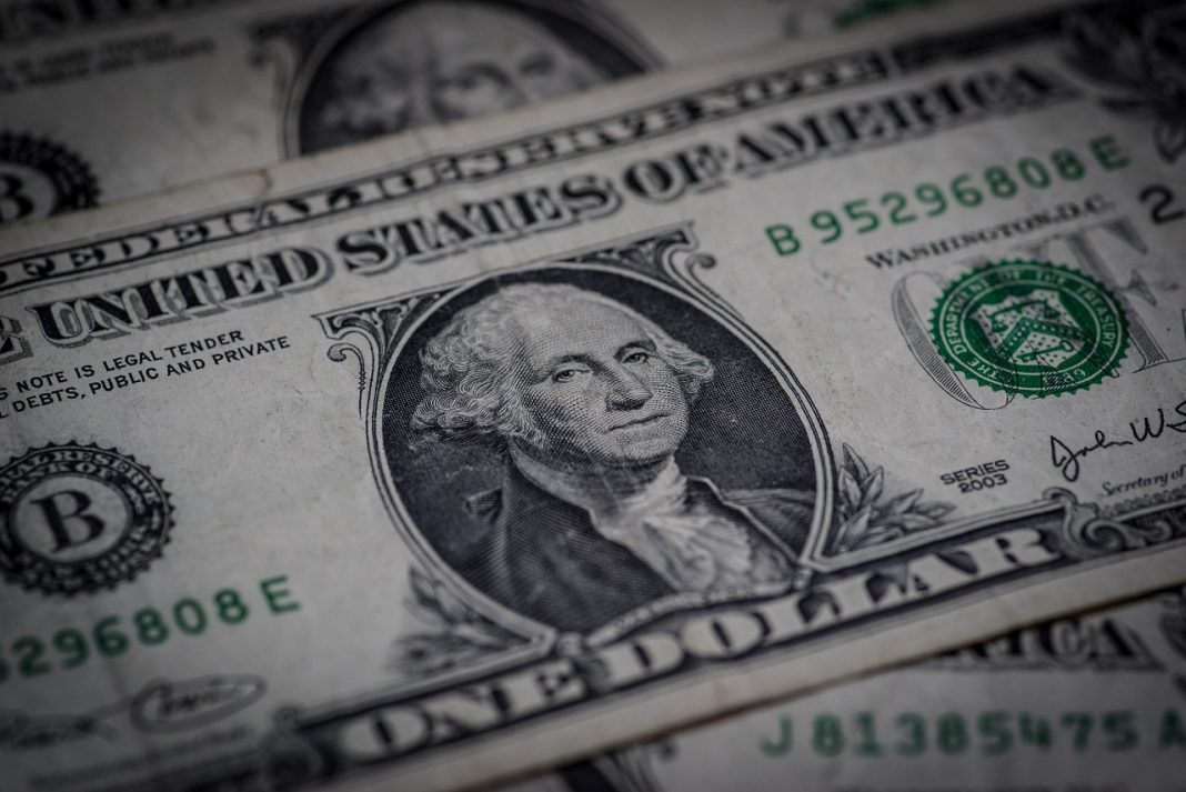 Nota de um dólar norte-americano em grande plano, com o retrato de George Washington ao centro e outras notas desfocadas em segundo plano.