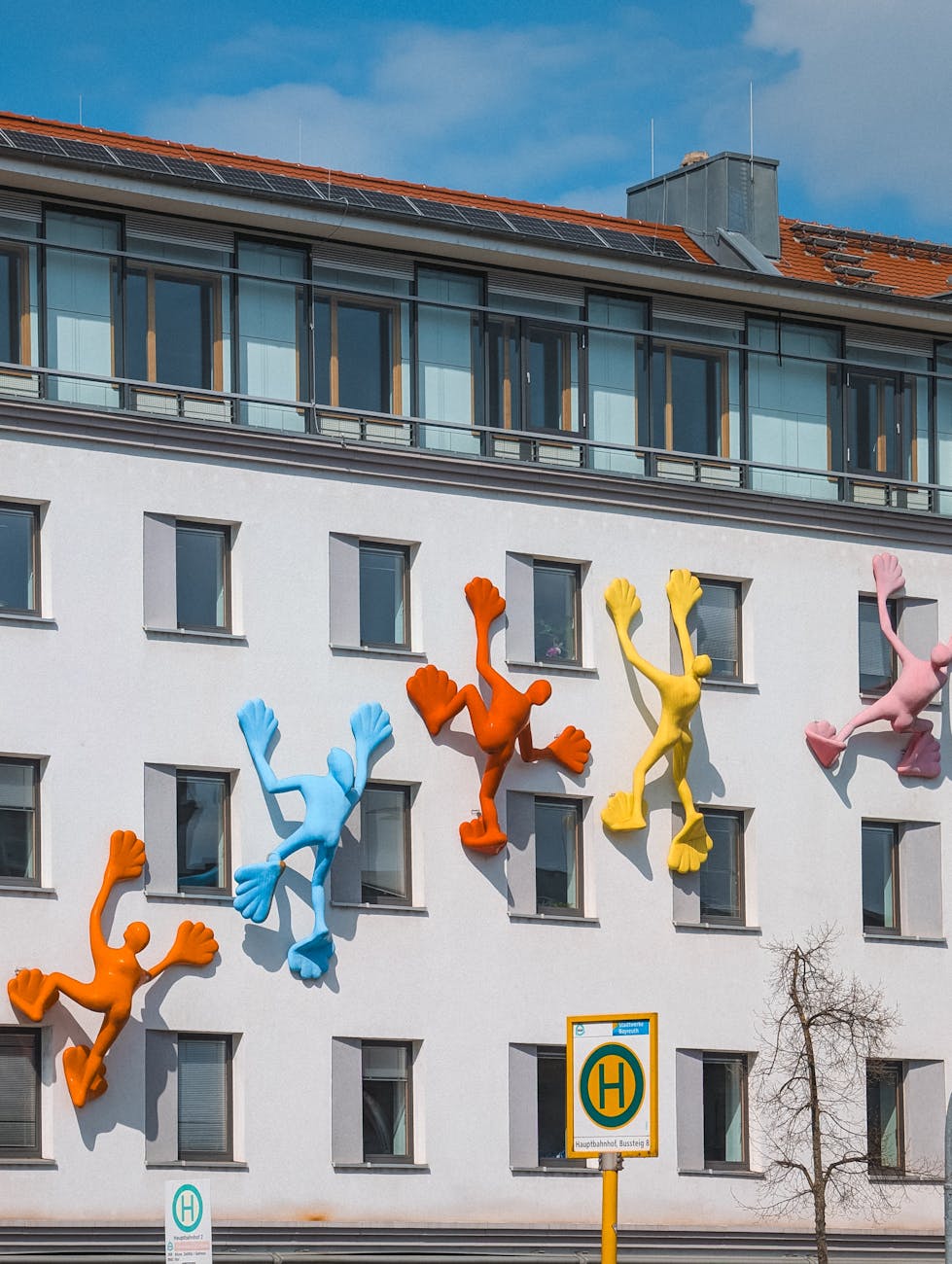 Figuras humanas coloridas fixadas numa fachada branca parecem escalar o edifício entre várias janelas.