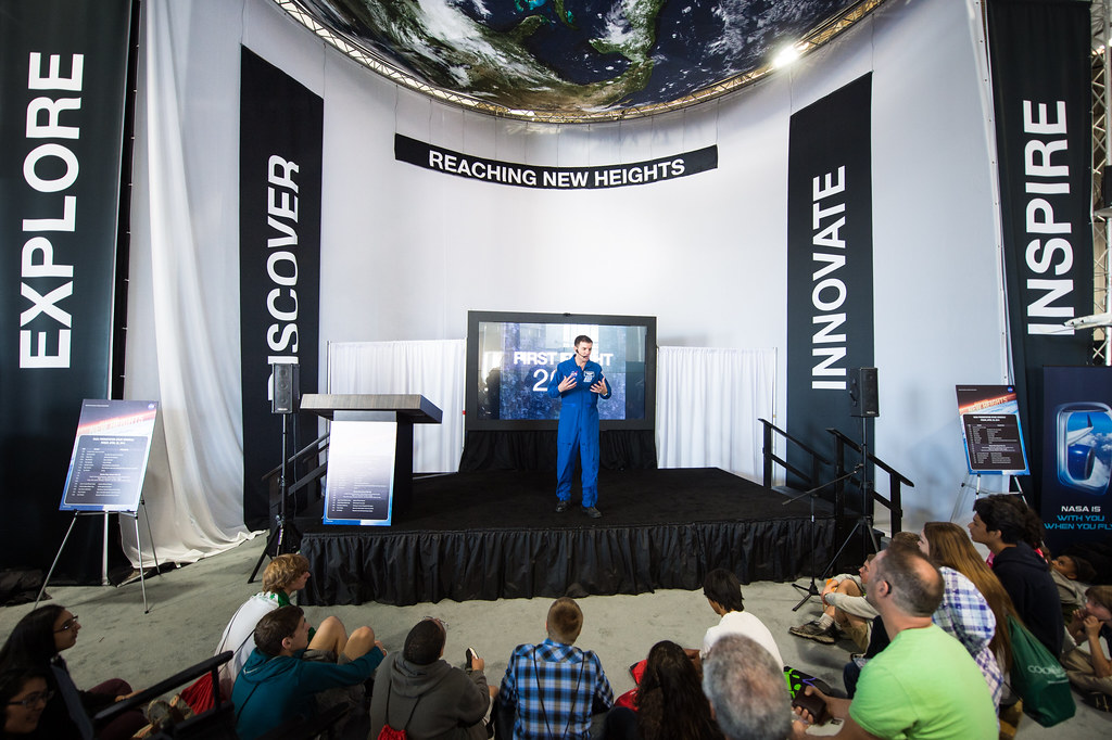 USA Science and Engineering Festival 2014 (201404250025HQ) |||| Orador em palco durante uma apresentação sobre exploração aeroespacial, com público sentado e painéis com palavras como explore, discover, innovate e inspire.