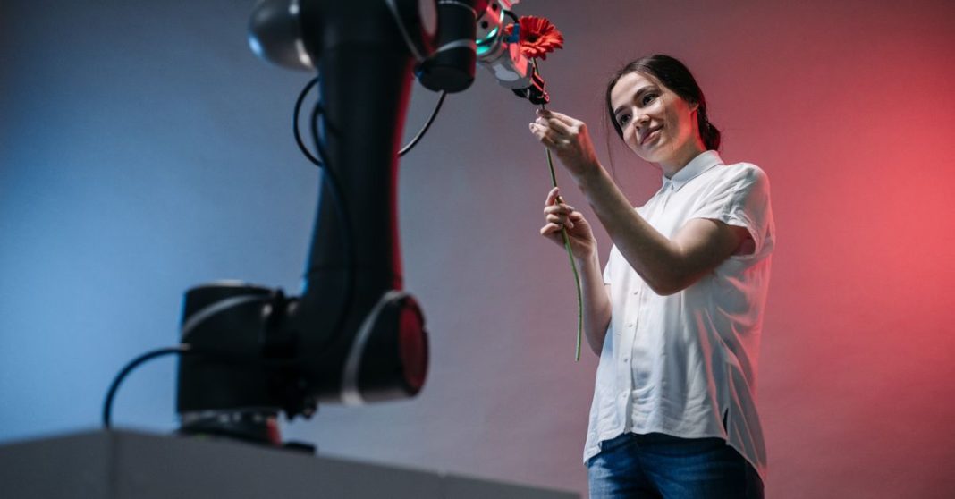 Mulher a interagir com braço robótico que segura uma flor, simbolizando a inteligência artificial, robótica e infraestrutura física.