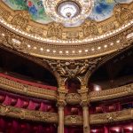 Interior ornamentado de um teatro clássico, com balcões dourados, camarotes vermelhos e teto decorado.
