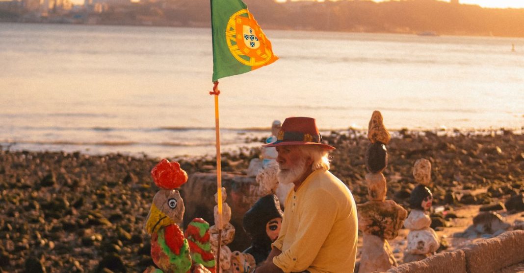Homem idoso junto à bandeira portuguesa, numa imagem editorial sobre pobreza, envelhecimento e falta de convergência social em Portugal.