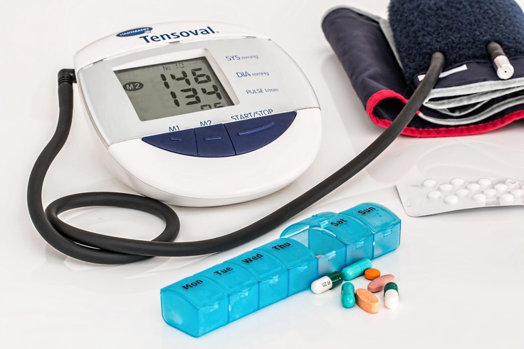Tensiómetro digital com braçadeira, organizador semanal de medicamentos e comprimidos sobre uma superfície branca.