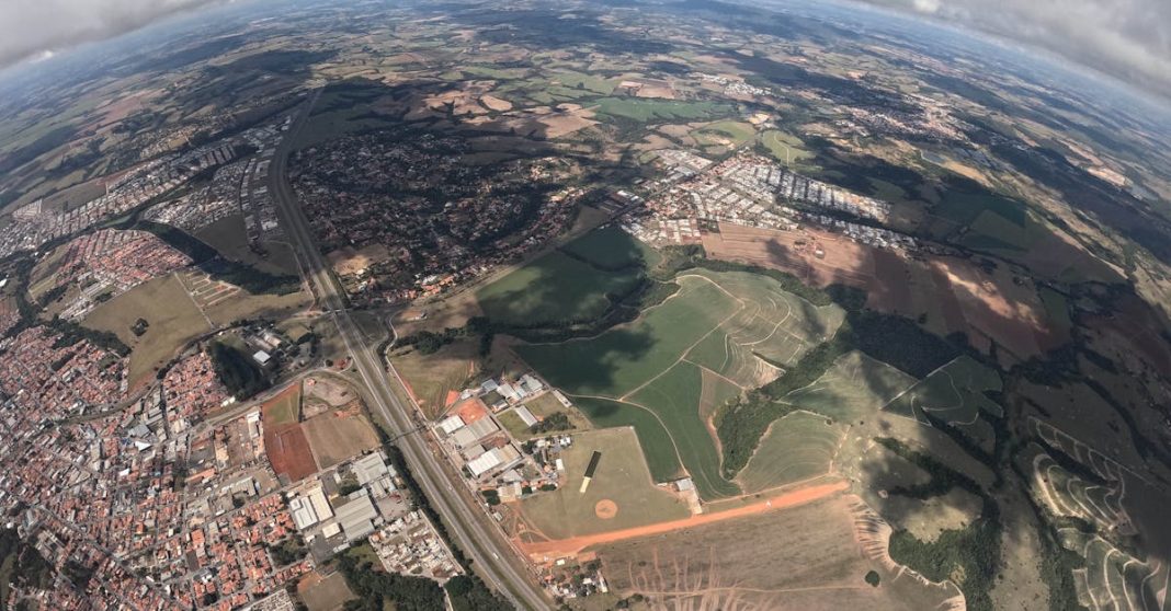 Vista aérea panorâmica de uma zona urbana e agrícola, com estradas, campos, nuvens e horizonte curvo visto a partir de grande altitude.