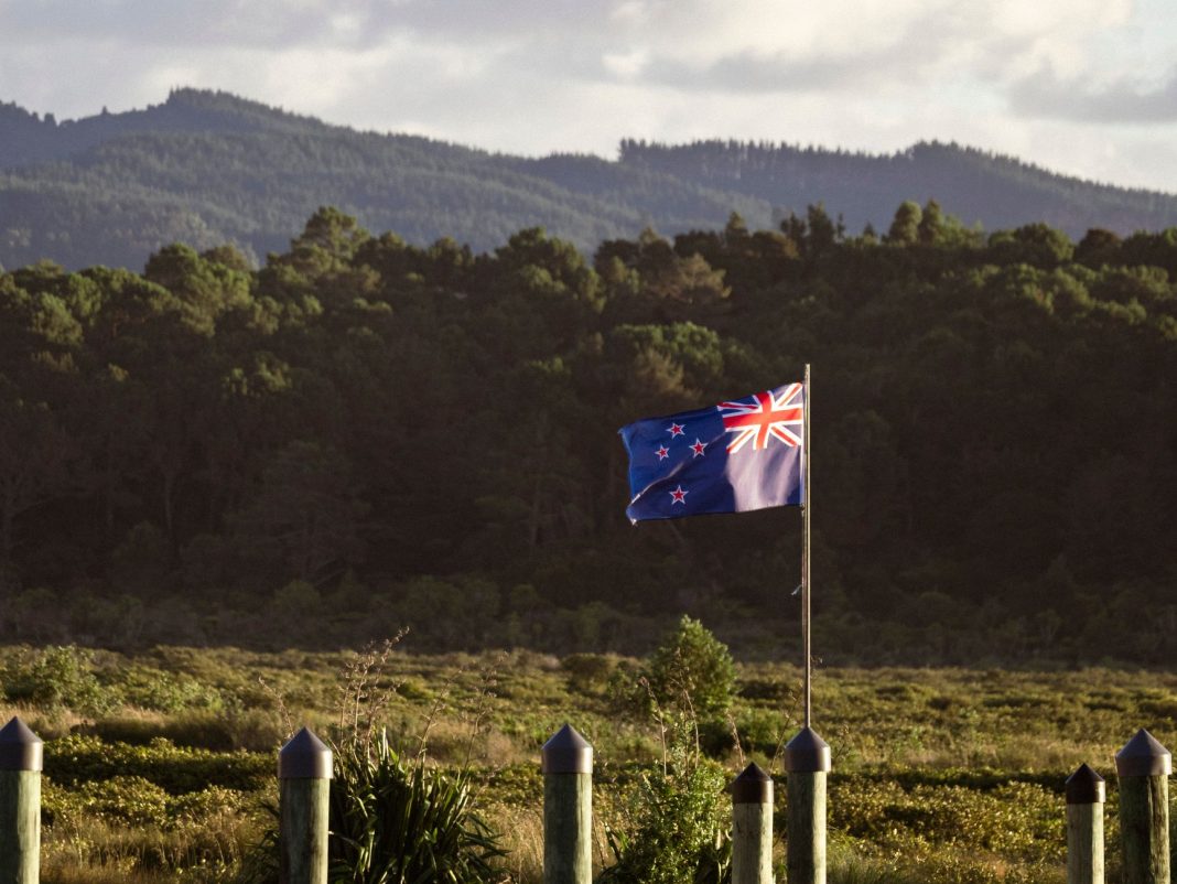 Bandeira da Nova Zelândia hasteada diante de uma paisagem rural e montanhosa.