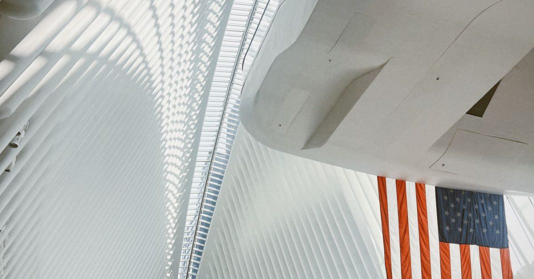 Bandeira dos Estados Unidos suspensa no interior de um edifício de arquitetura branca e monumental, com linhas verticais e luz natural