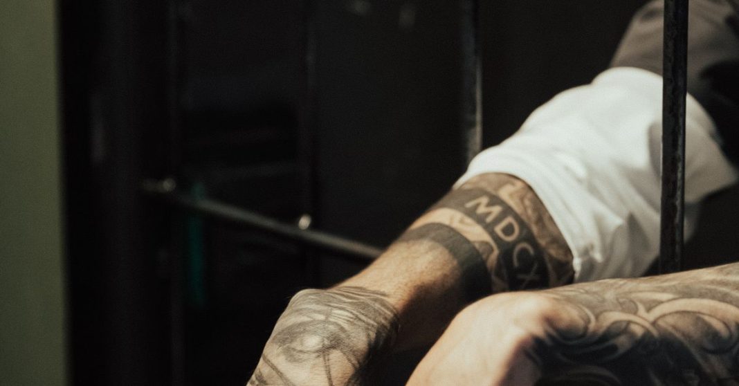 Close-up de um braço com tatuagens visíveis, apoiado entre as barras de uma cela, em ambiente de baixa luminosidade