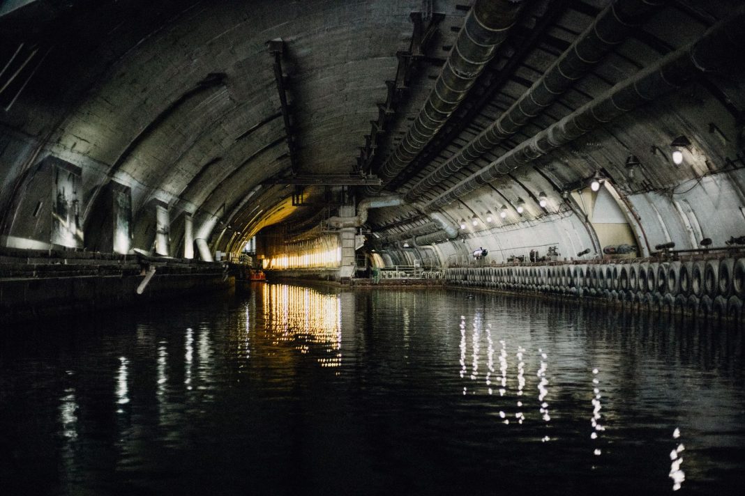 A Austrália Pode Ficar Sem os Submarinos Americanos | Interior de uma doca naval subterrânea, com canal de água, paredes de betão, tubagens e sistemas de ventilação.