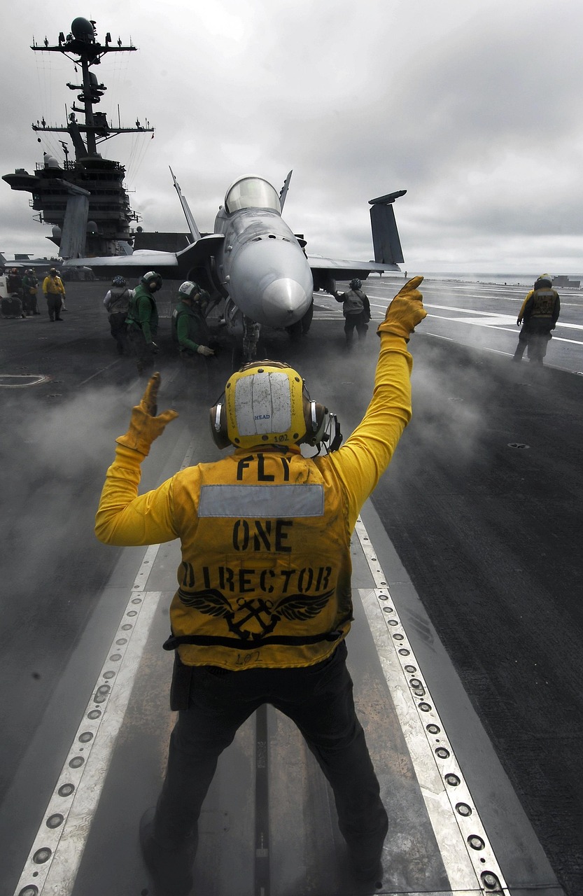 Marinheiro de fato amarelo faz sinais manuais a um caça F/A-18 no convés de um porta-aviões da Marinha dos EUA, rodeado de tripulantes, em condições de neblina.